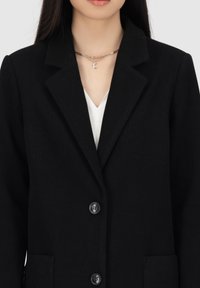 Zwarte blazer van een gestructureerde stof, met een reverskraag, knopen aan de voorkant en een enkele zak. Draagt over een witte top met een zilveren ketting.