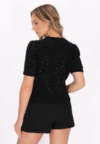 Pulover negru tricotat cu m�âneci scurte tip puff, având un design texturat și strasuri împrăștiate, cu tiv unde, purtat cu pantaloni scurți negri.