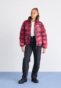 Pufová bunda v červené a černé gradientní barvě s vysokým límcem, zdobená žlutým logem, spárovaná s černými kalhotami a masivními černými botami.