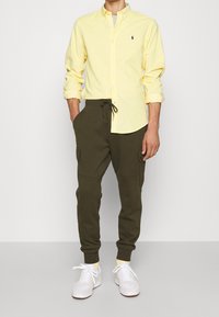 Světle žlutá košile s knoflíky, olivově zelené tepláky s kapsami a bílé tenisky. Ležérní outfit s volným střihem a stahovací šňůrkou.