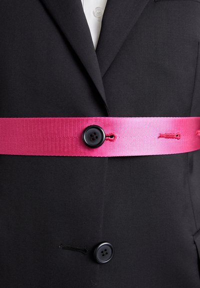 Veste noire avec une large ceinture en ruban rose, maintenue par deux boutons noirs. Le ruban a une finition texturée, ajoutant un contraste au tissu.