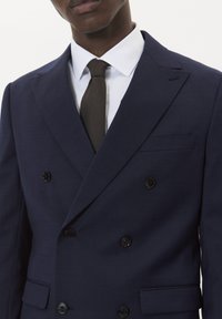 Mornarski sako na dvojno kopčanje od teksturirane tkanine s crnim gumbima. Bijela košulja i tamnosmeđa kravata upotpunjuju izgled.