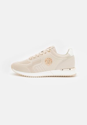 Beżowy dziany sneaker z wiązaniem z przodu, teksturowanym wzorem, okrągłym złotym logo po boku, białą podeszwą i metalicznym akcentem na pięcie.