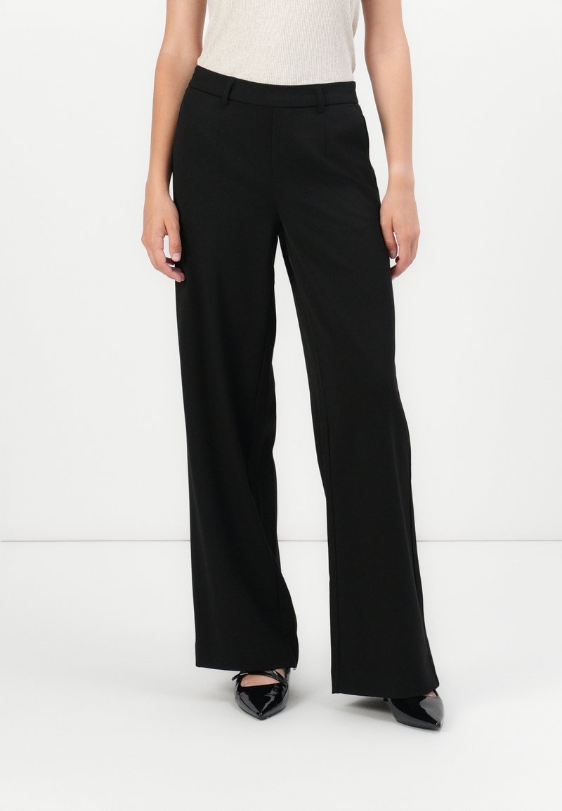 Femme portant un pantalon noir à jambes larges associé à un top beige sans manches et des chaussures noires à bouts pointus, debout sur un fond blanc.