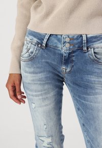 Personne portant un jean en denim bleu clair usé avec une fermeture à double bouton et un pull en maille beige côtelé, main détendue le long du corps.