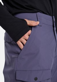Mano que lleva una camiseta de manga larga negra, con los dedos metidos en el bolsillo lateral de unos pantalones cargo morados con un bolsillo con tapa abotonada.
