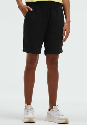 Person trägt schwarze Shorts mit umgeschlagenem Saum, gelbes Oberteil und weiße Turnschuhe und steht mit einer Hand in der Tasche vor einem einfachen Hintergrund.