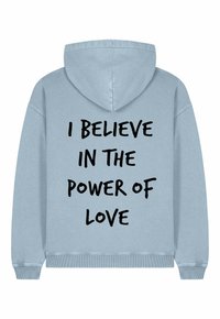 Felpa blu chiaro con cappuccio regolabile. Presenta una grande scritta nera sulla schiena: "CREDO NEL POTERE DELL'AMORE." Materiale in cotone, taglio casual.