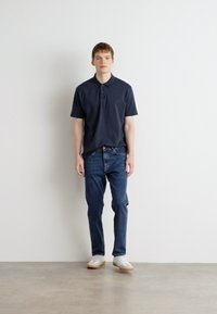 Cămașă polo bleumarin, blugi din denim și adidași albi. Cămașa are guler și mâneci scurte; blugii au o croială clasică. Elemente de design simple.