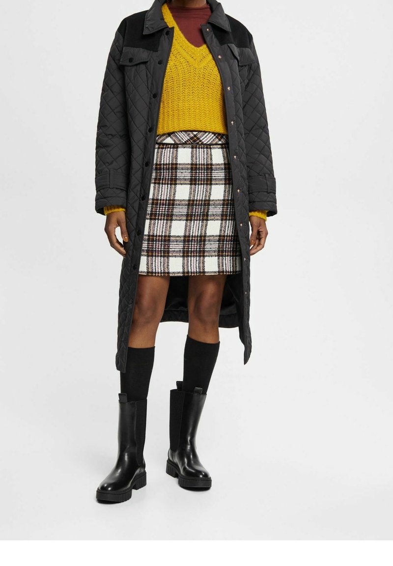 Abrigo largo acolchado negro, suéter de punto amarillo, falda de cuadros en blanco y negro, y botines negros de tacón grueso con calcetas hasta la rodilla.