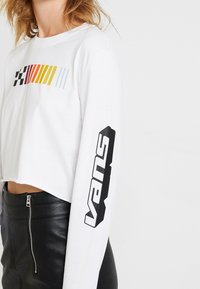 Vit kortärmad tröja med lång ärm och ett flerfärgat horisontellt randigt mönster samt svart "VANS"-logotyp på höger ärm.