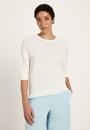 Young person with short curly hair wearing a white textured three-quarter sleeve top and light blue pants, standing with one hand in pocket.