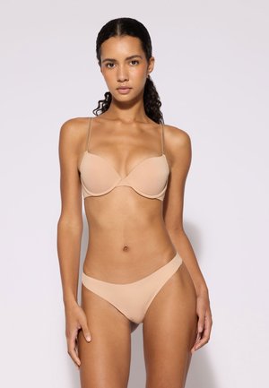 Donna con capelli scuri e ricci che indossa reggiseno color nudo e biancheria intima stile bikini, in piedi davanti a uno sfondo chiaro, rivolta in avanti.