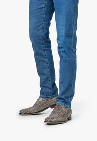 Stivali alla caviglia intrecciati in marrone e crema con punta affilata e tacco basso, abbinati a jeans in denim blu.