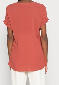 Camisetta ampia color ruggine con maniche arrotolate, realizzata in tessuto leggero, con scollatura rotonda e una sottile consistenza.