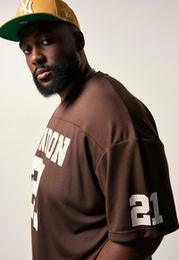 Homme avec une barbe portant un maillot de sport marron avec "21" sur la manche et une casquette jaune des New York Yankees, regardant la caméra.