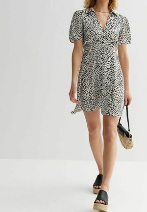 Mujer caminando con un vestido corto de manga corta blanco con manchas negras, sandalias negras de plataforma y sosteniendo un bolso de paja tejido con solapa negra.