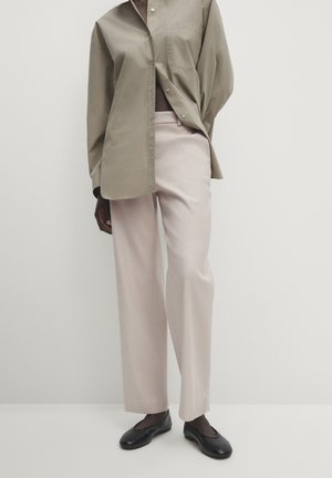 Osoba ubrana w beżową koszulę na guziki, jasnoróżowe szerokie spodnie i czarne płaskie buty, stojąca na tle jednolitego jasnego tła.