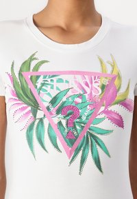T-shirt blanc à manches courtes avec un graphique triangulaire rose représentant des feuilles tropicales, des accents pailletés, et le nom de la marque "GUESS."