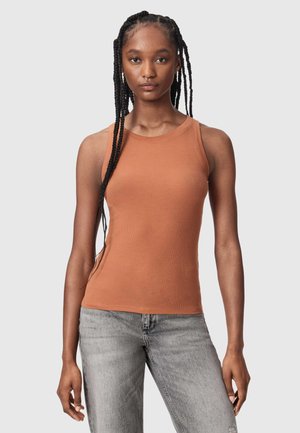 Jeune femme avec de longues tresses portant un haut côtelé sans manches couleur rouille-orange et un jean gris clair, debout devant un fond uni.