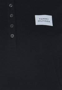 Polo noir avec quatre boutons foncés et un logo en patch blanc portant l'inscription "Tommy Hilfiger" sur la poitrine gauche.