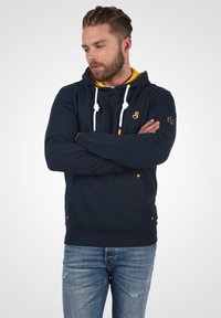 Mørkeblå hettegenser med gule detaljer og snorer, med en frontlomme i kengurostil og logo på brystet. Kombinert med lyseblå slitte jeans.
