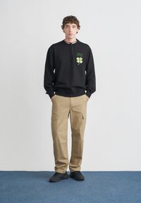 Felpa nera con stampa grafica verde, pantaloni cargo color cachi con tasche laterali e scarpe da ginnastica nere. Il modello è in piedi contro uno sfondo chiaro.