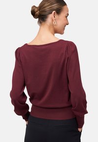 Donna con i capelli raccolti in un bun, che indossa orecchini a cerchio dorati, un top a maniche lunghe bordeaux e pantaloni neri, vista di spalle su uno sfondo bianco.