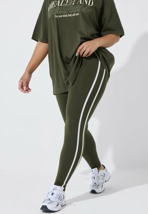 Vrouw die olijfgroene legging met witte zijstrepen draagt, bijpassend oversized T-shirt en witte sportschoenen, lopend op een witte achtergrond.