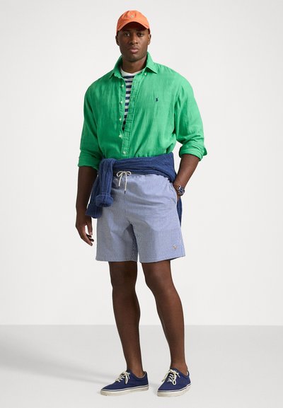 Chemise en lin verte, couche de dessous rayée, shorts rayés bleus, chaussures bleues marine et casquette orange. Un pull bleu marine noué autour de la taille ajoute du détail.