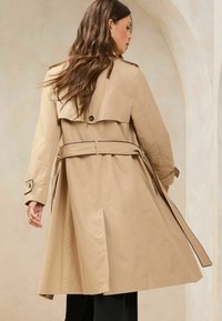 Beige trenszkkabát, dupla gombos kivitelben, övvel a deréknál, széles gallérral és gombos mandzsettákkal. Simább anyag, kontrasztos piping részletekkel.