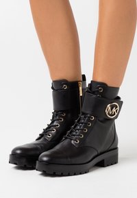 Botas de cuero negro hasta el tobillo con punta redonda, detalles de herrajes dorados, cremallera lateral y un puño ancho con un detalle de logo. Suela texturizada.