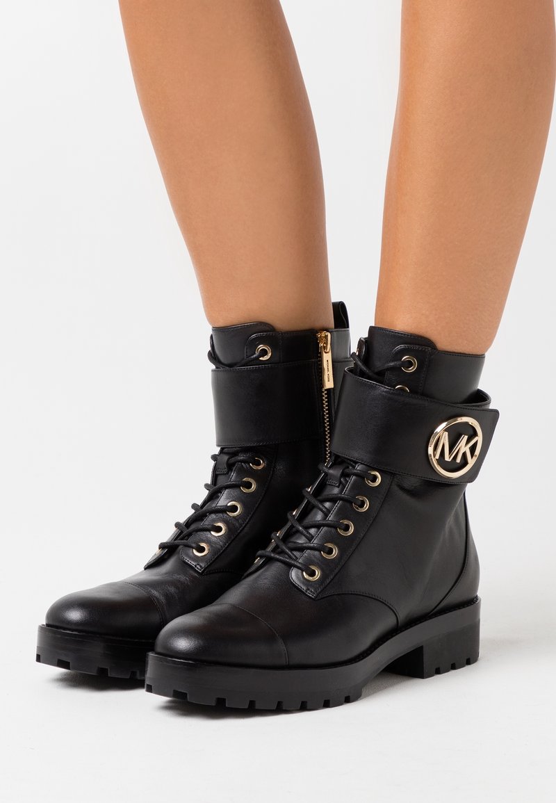 Botas de cuero negro hasta el tobillo con punta redonda, detalles de herrajes dorados, cremallera lateral y un puño ancho con un detalle de logo. Suela texturizada.