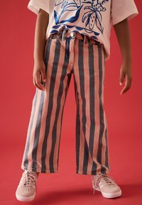 Los pantalones de mezclilla a rayas presentan rayas verticales en tonos rosa y azul oscuro, con un ajuste holgado y un cierre frontal de botones, combinados con zapatillas ligeras de color rosa.