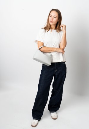 Srebrna usnjena torbica s strukturirano obliko in kratkim ročajem, v kombinaciji z belo majico in temnimi širokimi hlačami iz denima na nevtralni podlagi.