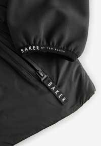 Sort jakke med en glat overflade, sammenkrøllede ærmekanter og en lynlås med en mærkeører. Teksten siger "BAKER by TED BAKER."