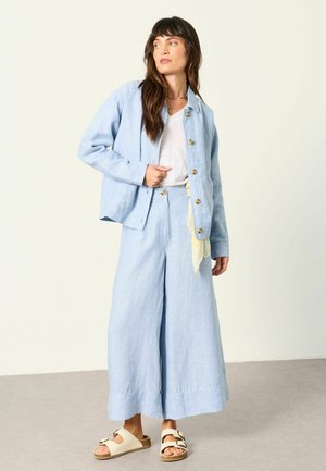 Kobieta ubrana w jasnoniebieską zapinaną na guziki kurtkę i szerokie spodnie, na białej koszuli, w beżowych sandałach i z naszyjnikiem, stojąca w pomieszczeniu.