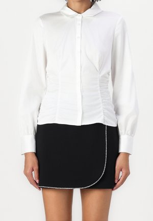 Femme portant une chemise blanche à manches longues avec des côtés froncés et une mini-jupe noire avec un détail chaîne sur l'ourlet enveloppant.