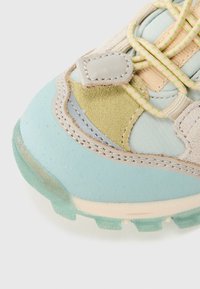 Κοντινό πλάνο στην μύτη ενός αθλητικού παπουτσιού με μπλε λαστιχένια σόλα, μπεζ σουέτ, ανοιχτοπράσινο ύφασμα και κορδόνια με πουά σε νεον πράσινο χρώμα.