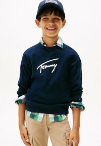 Ragazzino sorridente che indossa una felpa navy blue di Tommy, una camicia a quadri verde, pantaloni cargo beige e un cappellino navy di Tommy su uno sfondo bianco.