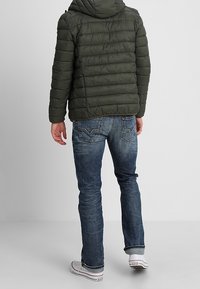 Doudoune verte à coutures horizontales, avec capuche, portée avec un jean bleu délavé et des baskets blanches, vue de dos.