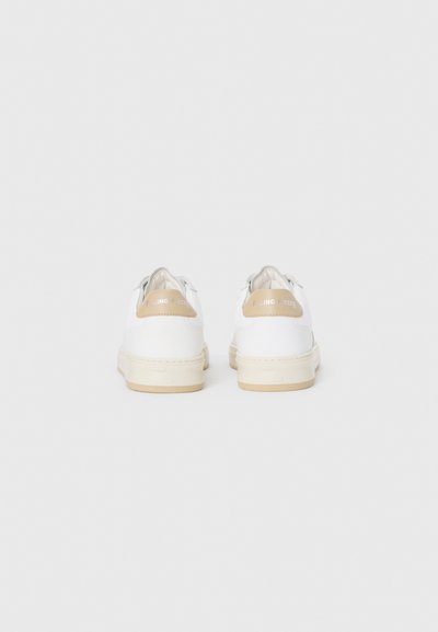 Et par hvite lave sneakers med beige hæltabber og off-white såler, sett fra baksiden på en ensfarget bakgrunn.
