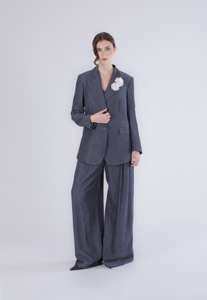 Kvinde iført en oversized mørkegrå dragt med bukser med vide ben og en hvid blomsterbroche, stående foran en ensfarvet lys baggrund.