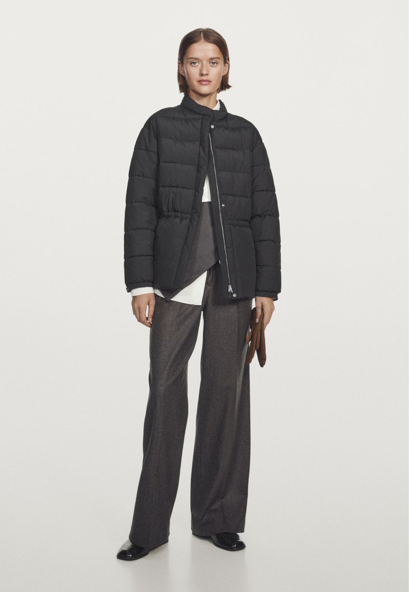 Giubbotto imbottito nero con colletto alto, chiusura con zip e design trapuntato. Il modello indossa pantaloni marroni e tiene in mano dei guanti marroni, su uno sfondo bianco.