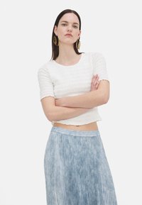 Hvit kortermet topp med teksturert stoff, cropped design og scallop-kant. Kombinert med et lyseblått plissert skjørt. Gylne hoop-øreringer.