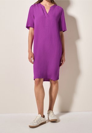 Femme portant une robe violette mi-longue à manches courtes avec un décolleté en V et des baskets blanches, debout contre un mur beige.