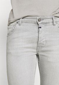 Jeans de denim gris claro con corte ajustado, que cuentan con bolsillos frontales, cremallera y cierre de botón metálico. Detalles sutiles de desgastado y costura.