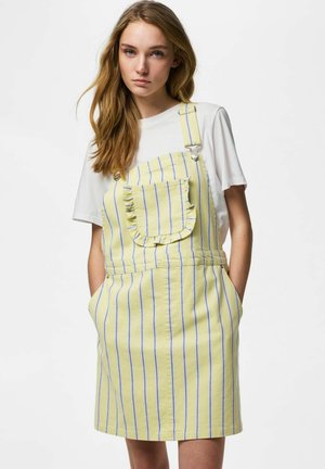 Ung kvinne med langt hår iført gul og blå stripete forkle-kjole over en hvit t-skjorte med korte ermer, hender i lommer, nøytral bakgrunn.