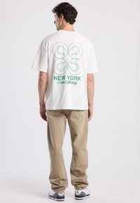 Weißes Baumwoll-T-Shirt mit grünem grafischem Druck eines Schleifenmotivs und dem Text "NEW YORK Tennis Society" auf dem Rücken, kombiniert mit beigen Hosen.