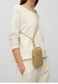 Borsa a tracolla beige con una texture imbottita, forma rettangolare e tracolla intrecciata, indossata sopra una felpa color crema e pantaloni della tuta.